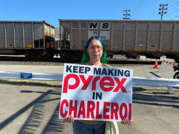 Erin Guzik holds a sign "KEEP MAKING PYREX IN CHARLEROI" as a train passes behind her.